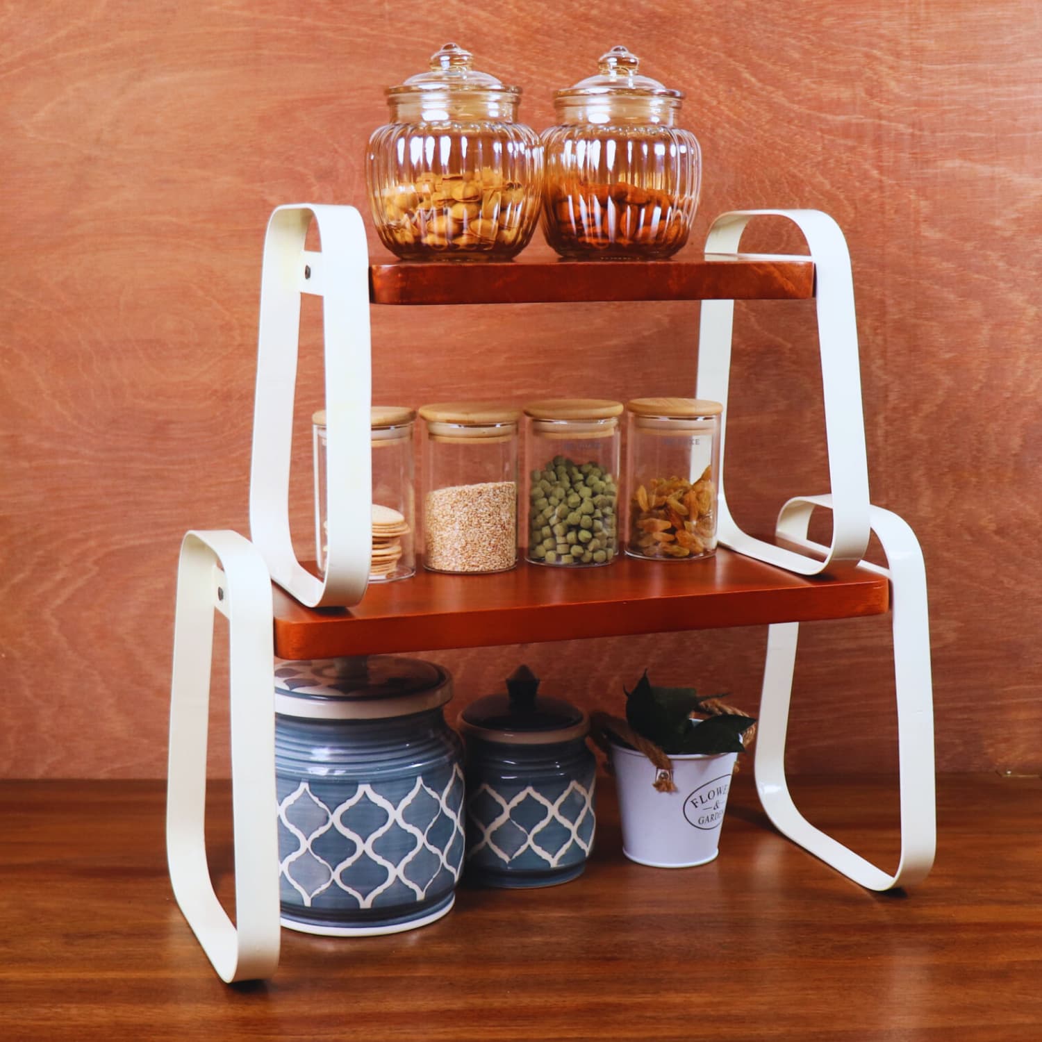 A two-tier wooden shelf with white metal supports holds various kitchen items. The top tier has two glass jars with snacks, the middle tier has three spice containers, and the bottom holds ceramic canisters and a small potted plant. Buy LOOSEBUCKET's The Stackable Wooden Kitchen Organizer Set of 2 online for an elegant touch to your space.