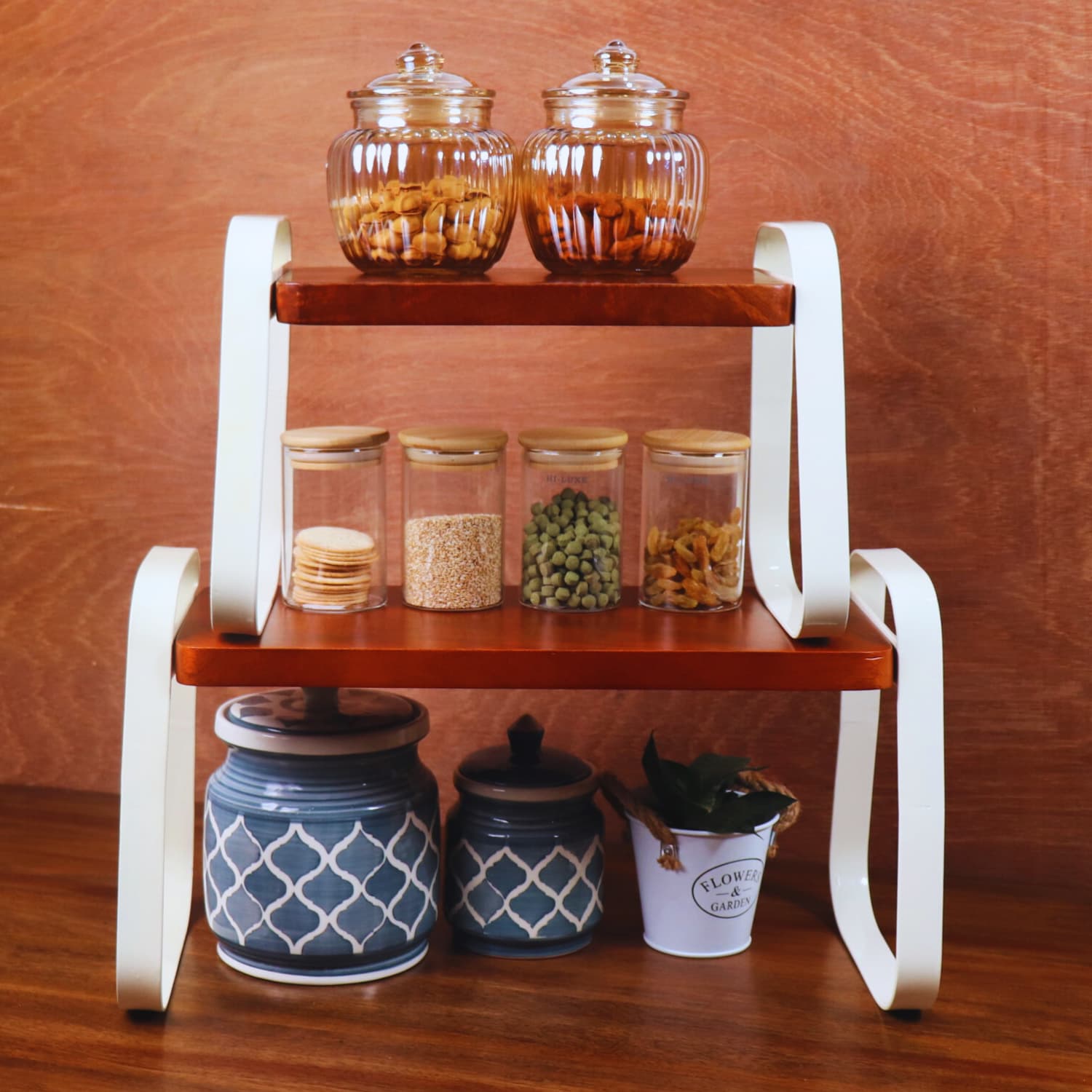 A wooden kitchen shelf with three tiers holds various items: the top tier has two glass jars with nuts, the middle tier has three glass jars containing different food items, and the bottom tier has two blue ceramic containers and a small plant in a white pot. Buy LOOSEBUCKET's best "The Stackable Wooden Kitchen Organizer Set of 2" online today!