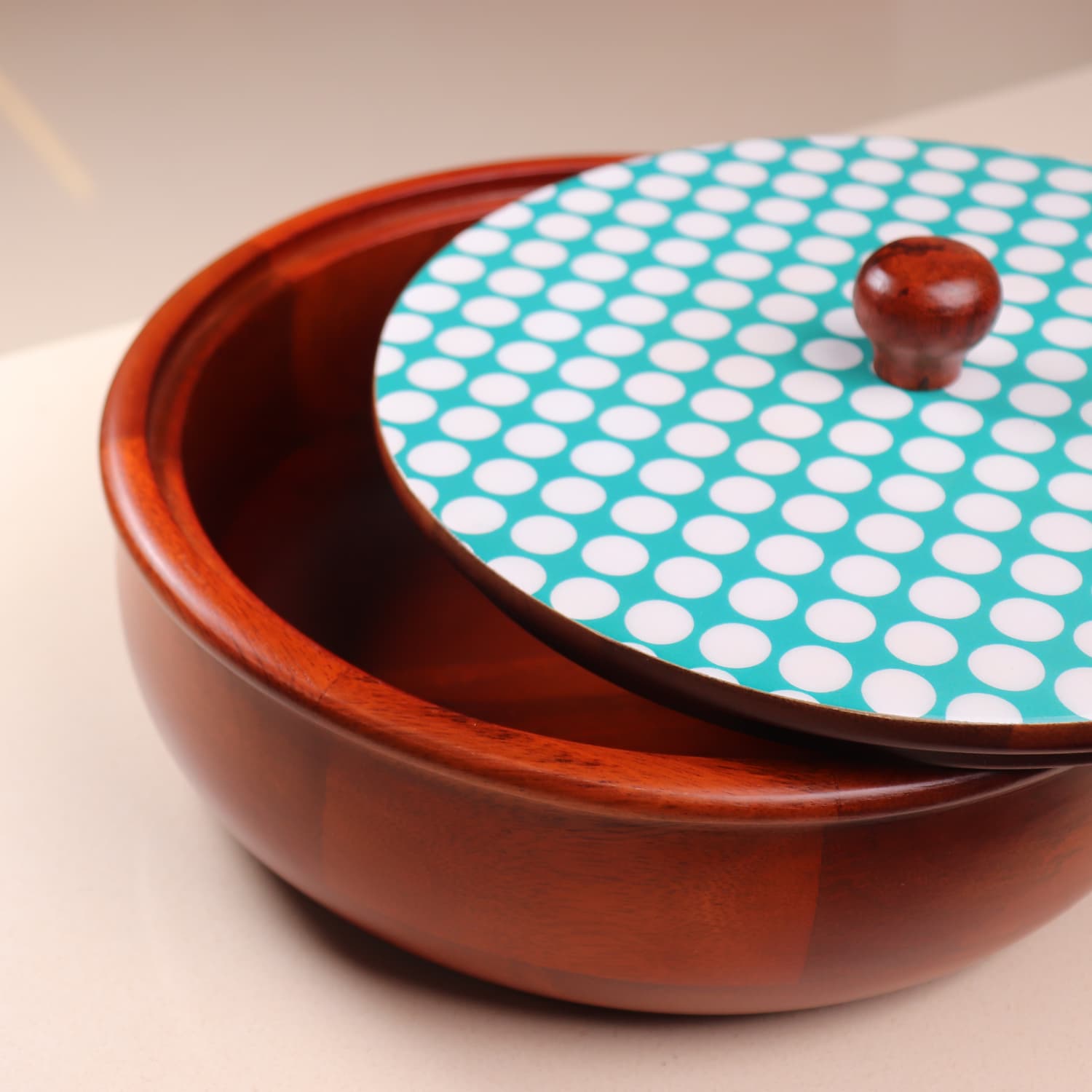 A premium wooden Roti Box with a slightly open matching lid. The lid has a turquoise background with white polka dots and a wooden knob handle. This elegant piece, often regarded as the best wooden Chapati box in India, rests on a smooth, light-colored surface. 

Introducing The Dot Legacy Chapati Box by LOOSEBUCKET, which brings both functionality and style to your kitchen.