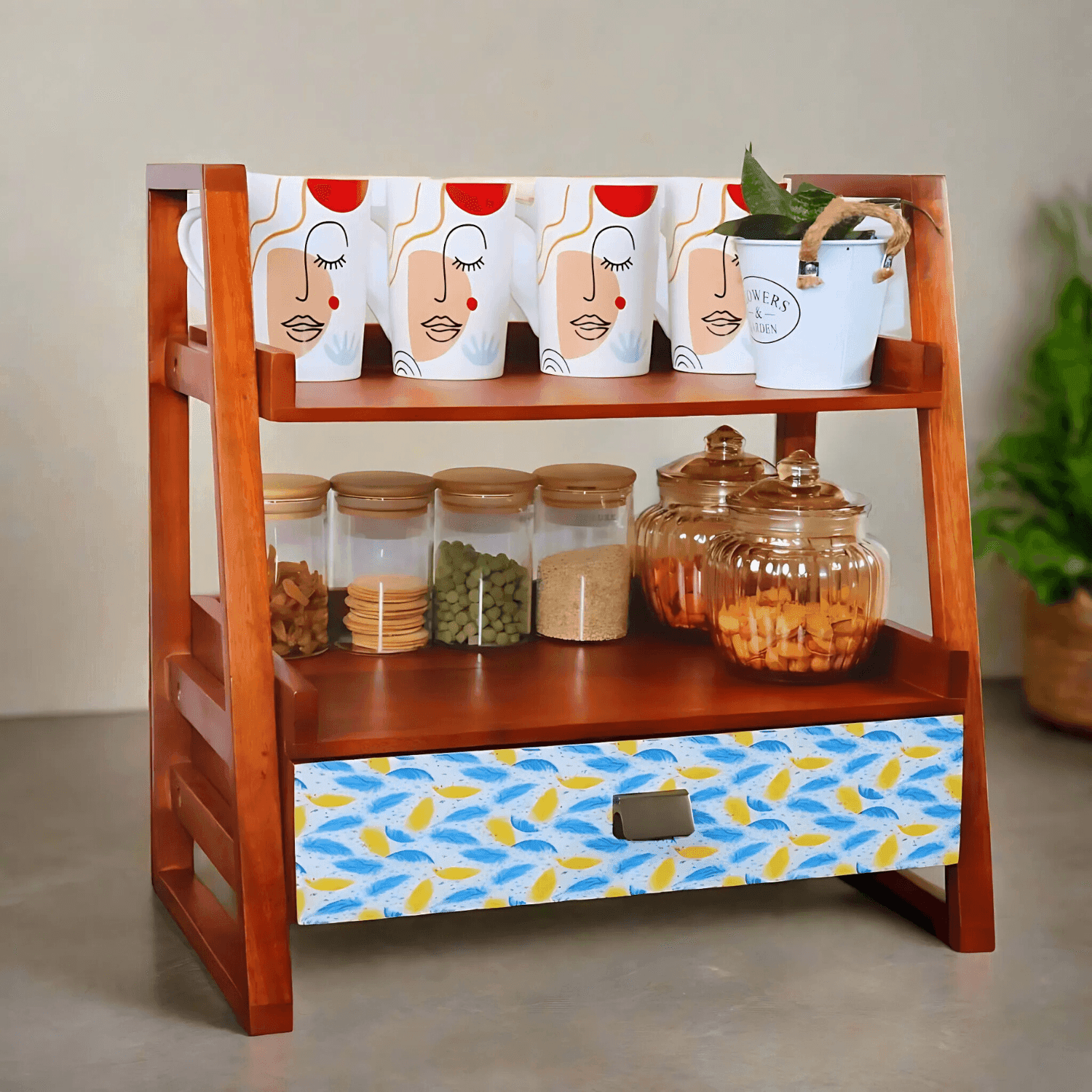 The Whispering Feather wooden kitchen organizer featuring two-tiered walnut brown shelving with decorative mugs, glass storage jars, and a blue-yellow feather pattern drawer. Handcrafted mango wood construction ideal for kitchen storage and display.