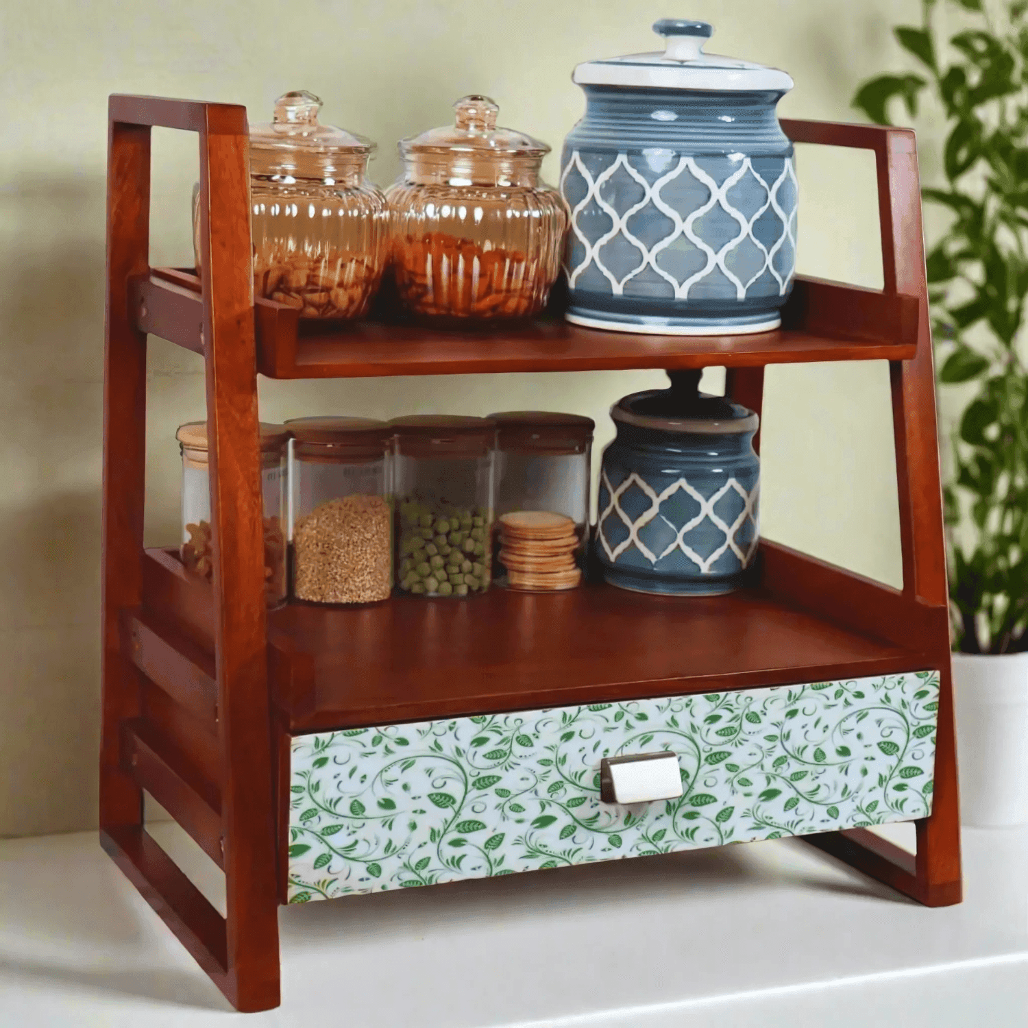 The Greenvibes Wooden Kitchen Versatile Organizer featuring walnut-brown mango wood frame with two shelves displaying glass storage jars and blue ceramic containers, complemented by a decorative green floral pattern drawer at the bottom.