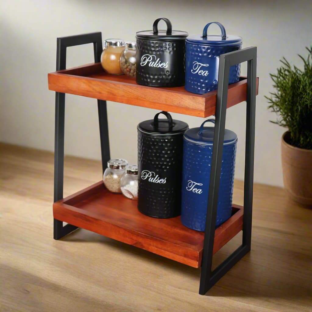 Loosebucket Linea Premium Wooden Kitchen Organizer featuring two-tier walnut-finished acacia wood shelves with black metal frame, displaying decorative blue and black storage canisters for tea and pulses, with glass jars on wooden surfaces. Small potted plant visible beside the organizer.